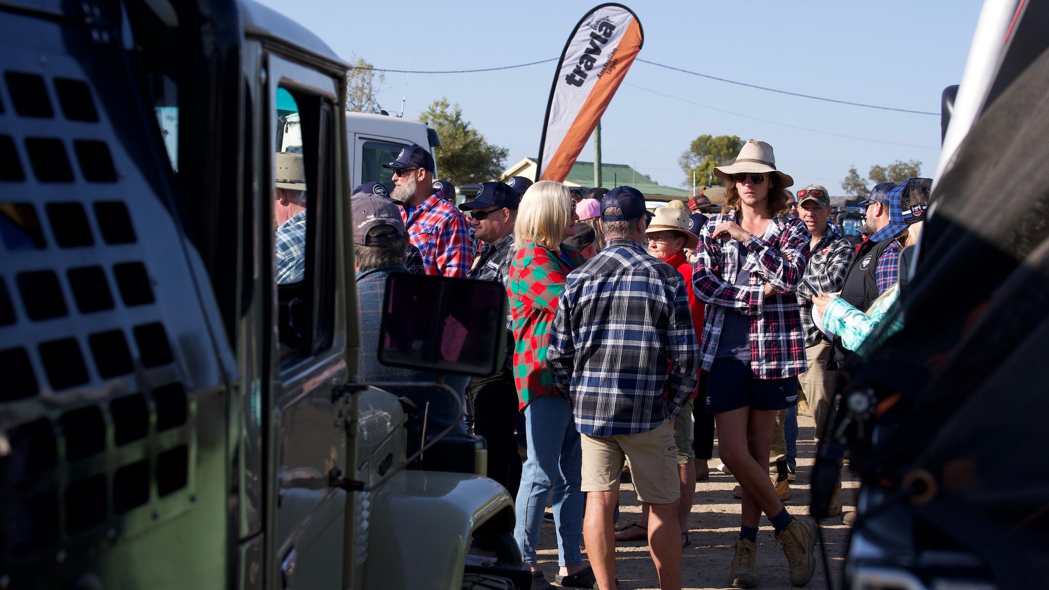 Crowd of farmers in flannos next to 4wd vehicles, travla flag in the background